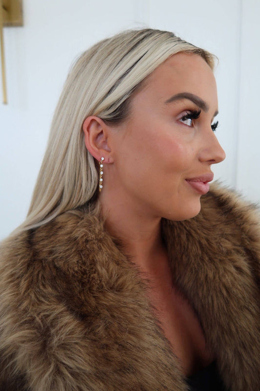 Woman wearing a fur coat with a neutral background, wearing gold/diamond drop earrings
