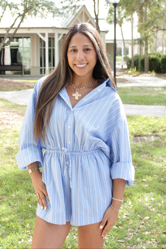 Woman in a blue striped shirt romper standing outdoors with trees and a building in the background