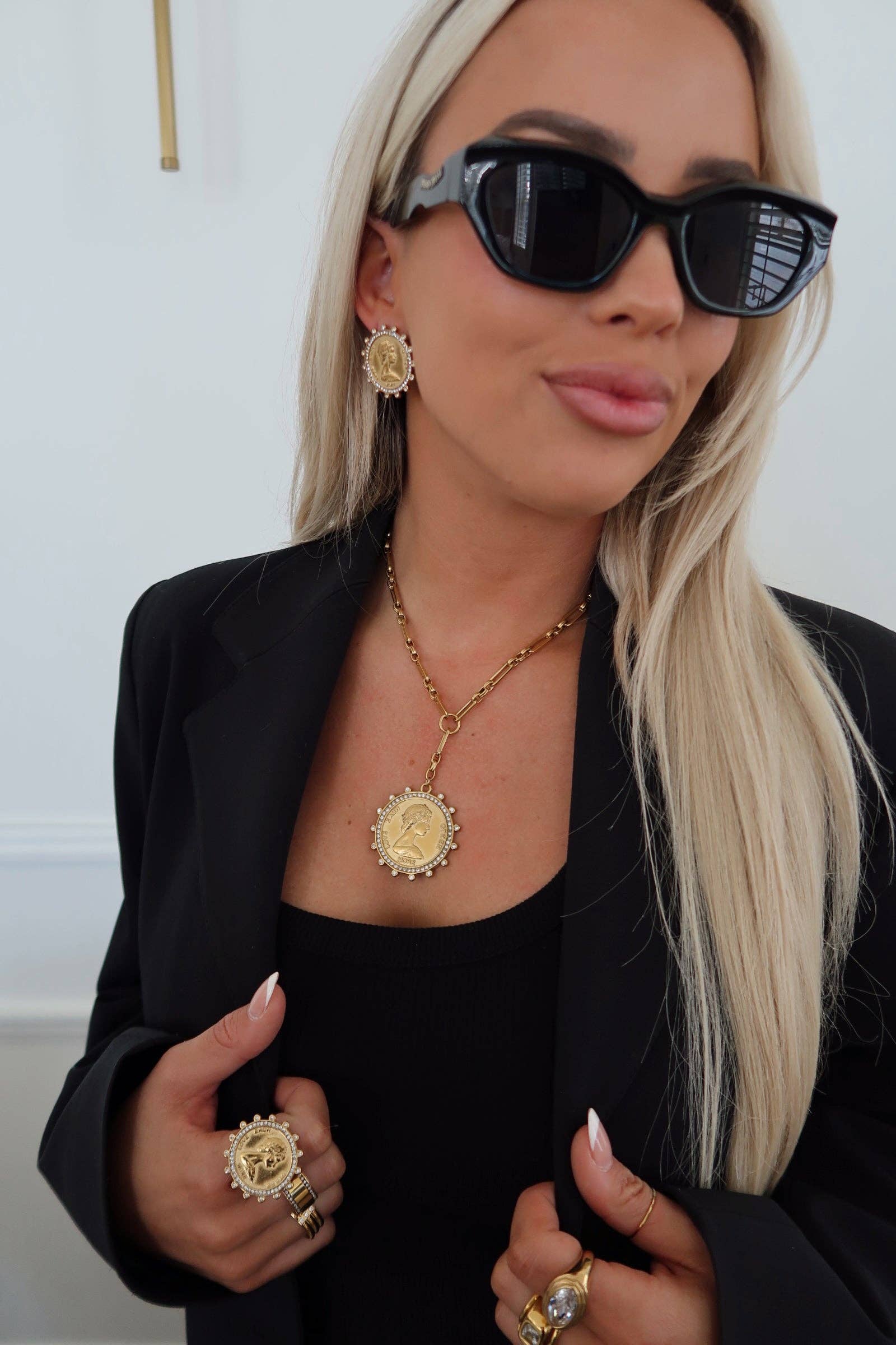 Woman wearing sunglasses, black blazer, and gold jewelry indoors.