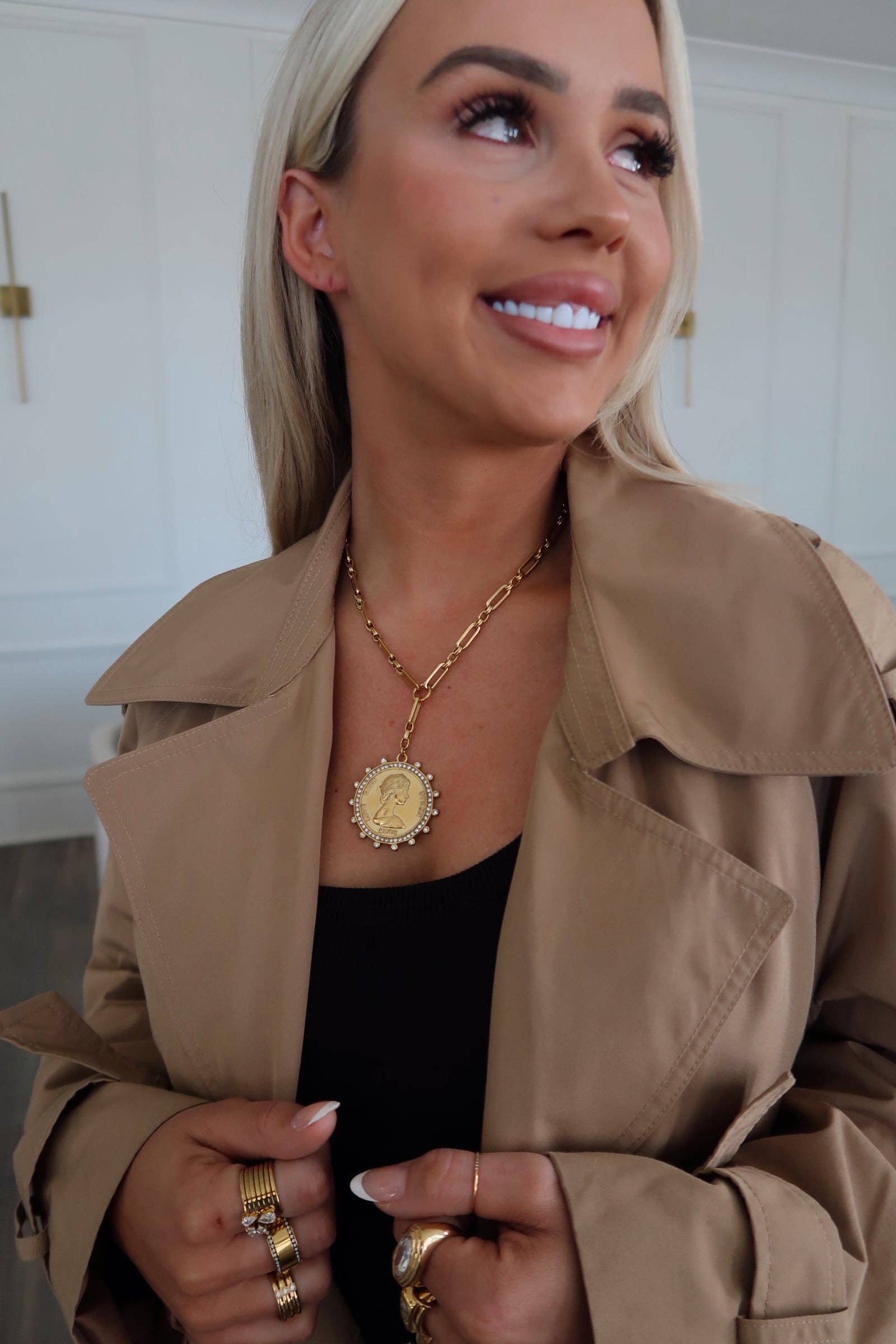 Woman wearing a beige trench coat and gold jewelry indoors.
